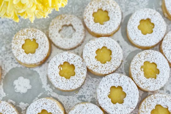 Baked Lemon Curd Linzer Cookies placed on white surface dusted with powdered sugar. In the top right corner is visible small part of yellow flower.