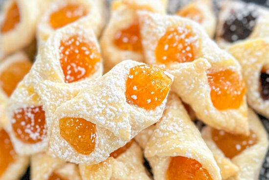 Baked Kolaczki cookies stacked on top of each other and placed on white parchment paper, dusted with powdered sugar.