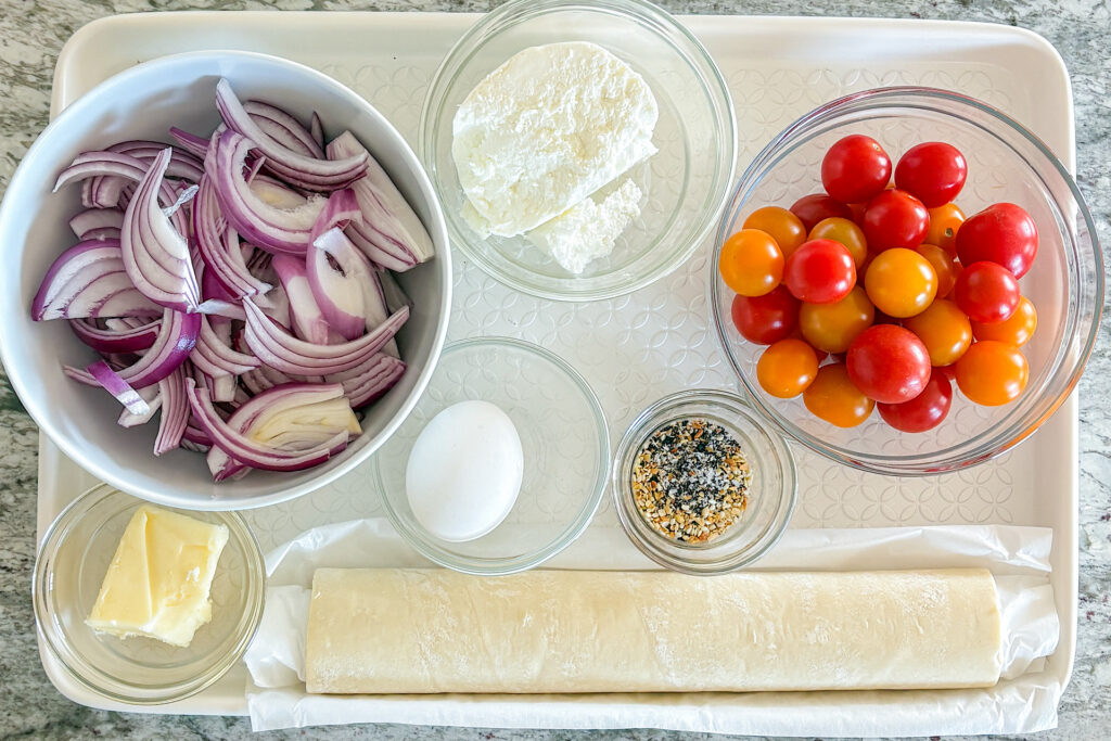 Ingredients for cherry tomato goat cheese tarts are arranged on a light beige baking sheet. In a white ceramic bowl are thinly sliced red onions. Next to it is a small glass bowl filled with goat cheese, followed by a larger glass bowl with yellow and red cherry tomatoes. Nearby is a small glass bowl of everything bagel seasoning, another small glass bowl with one egg, and a small glass bowl with butter. A sheet of puff pastry is also placed alongside the ingredients. The baking sheet is set on a light grey marble countertop.