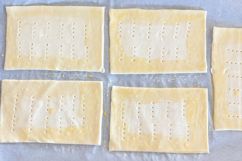 Four rectangular puff pastry pieces are fully visible, with half of a fifth piece showing on the right side. The pastry edges are brushed with egg wash, and the centers are pricked with a fork. Everything is placed on white parchment paper.