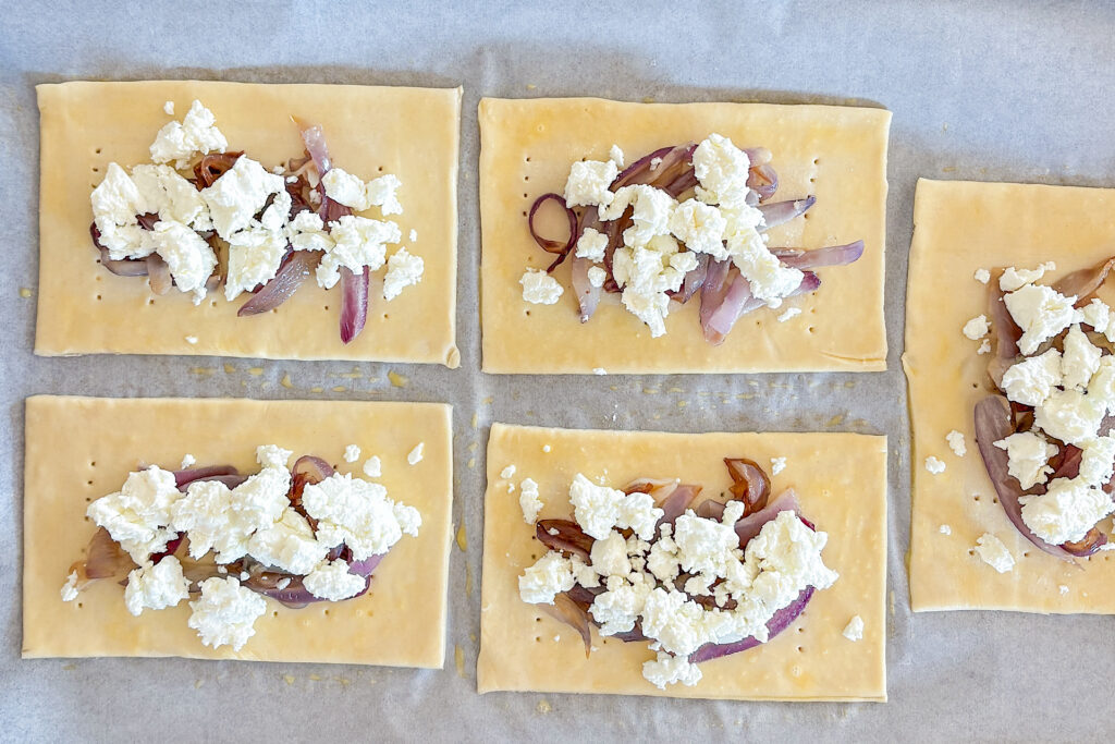Four rectangular puff pastry pieces are fully visible, with half of a fifth piece showing on the right side. In the center of each piece is caramelized cooked red onion, topped with crumbled goat cheese. Everything is placed on white parchment paper.
