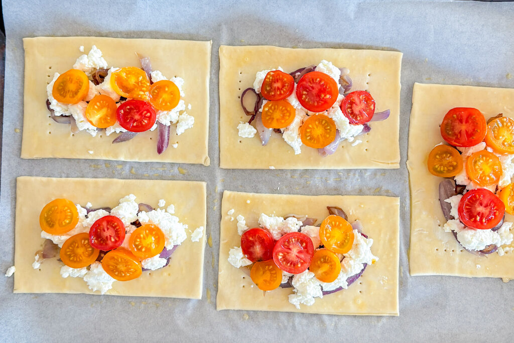 Four rectangular puff pastry pieces are fully visible, with half of a fifth piece showing on the right side. In the center of each piece is caramelized cooked red onion, topped with crumbled goat cheese, and on top of that are halved yellow and red cherry tomatoes. Everything is placed on white parchment paper.