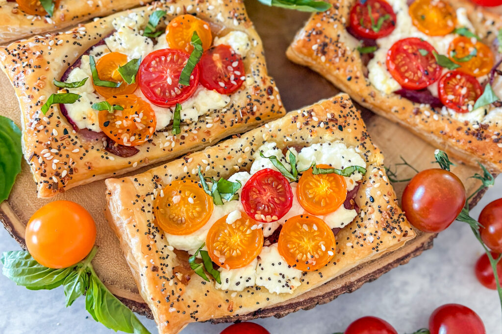 Two baked puff pastry pieces and one additional piece are placed on a round brown wooden board. In the bottom left corner, there is a yellow cherry tomato with fresh basil leaves. On the bottom right side, red cherry tomatoes are placed. Everything is arranged on a white surface.
