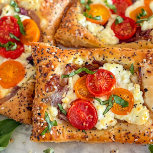 A baked puff pastry cherry tomato goat cheese tart topped with cherry tomatoes, goat cheese, and caramelized onions. The creamy melted goat cheese is layered with halved cherry tomatoes, finished with fresh basil and a sprinkle of everything bagel seasoning over the golden, flaky crust. Two similar tarts sit beside it. In the background, a few red cherry tomatoes are slightly visible in the top left corner. Fresh basil rests in the bottom left, and a single cherry tomato appears in the lower right. Everything is arranged on a white surface.
