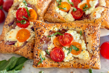 A baked puff pastry cherry tomato goat cheese tart topped with cherry tomatoes, goat cheese, and caramelized onions. The creamy melted goat cheese is layered with halved cherry tomatoes, finished with fresh basil and a sprinkle of everything bagel seasoning over the golden, flaky crust. Two similar tarts sit beside it. In the background, a few red cherry tomatoes are slightly visible in the top left corner. Fresh basil rests in the bottom left, and a single cherry tomato appears in the lower right. Everything is arranged on a white surface.