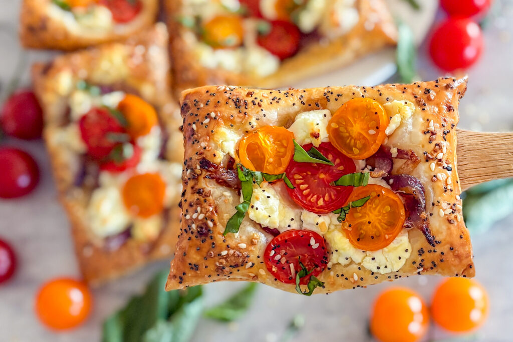 One baked puff pastry cherry tomato goat cheese tart is placed on a wooden spatula, held in the air. In the background, on a white surface, more baked tarts are visible, slightly blurred, along with fresh yellow and red cherry tomatoes beside them.