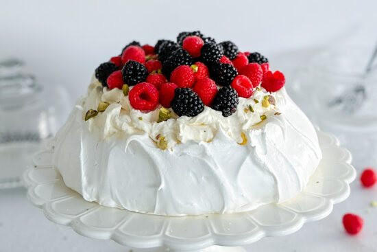 A baked Pavlova is topped with cream, fresh raspberries, blackberries, and sprinkled with crushed pistachios, sitting on a white porcelain cake stand. The stand rests on a clean white surface, surrounded by more raspberries and blackberries. In the background, a plate is partially visible, covered with a glass dome.