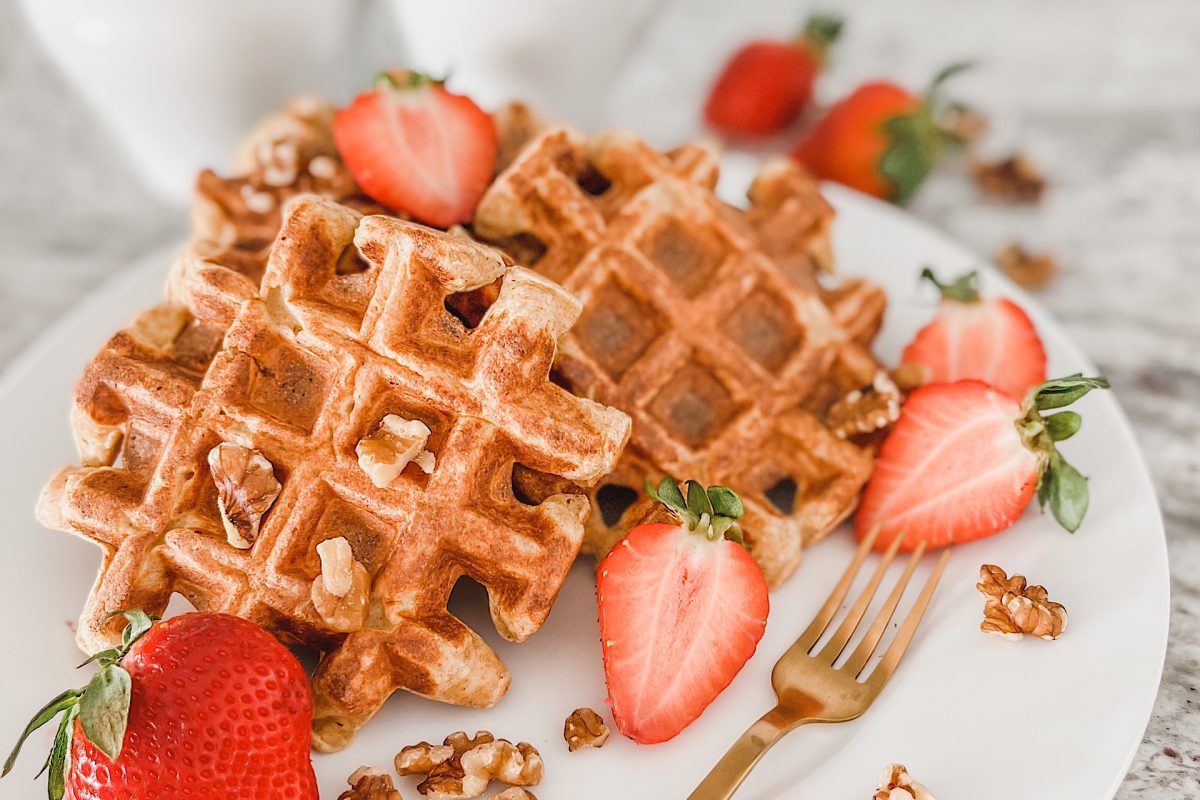 Lemon ricotta waffles with some halves of Strawberries and a few walnuts, on white porcelain plate, with small gold fork.