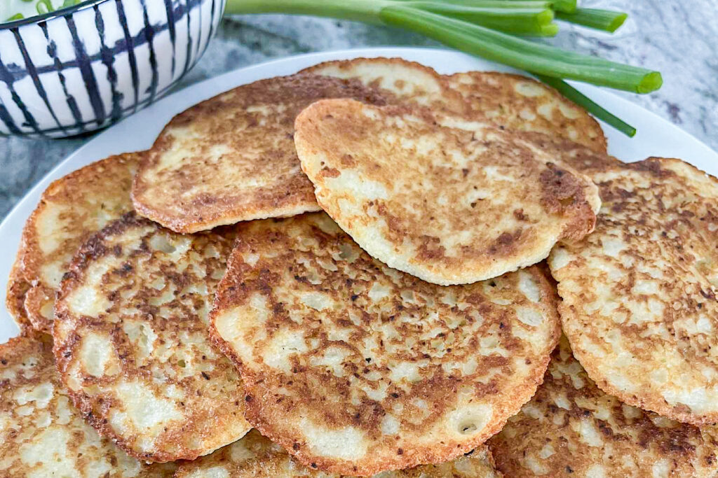 Fried crispy, tender Polish potato pancakes stack on a white plate. The plate is placed on a grey marble countertop. Beside the plate, on the left top side of the plate, is a small white serving bowl with black stripes filled with sour cream and topped with fresh green onions. Next to the bowl and plate is a bunch of fresh green onions.