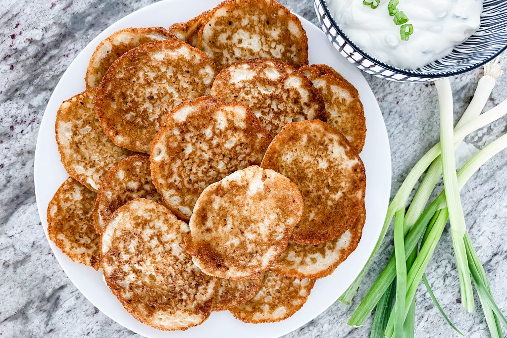 Fried crispy, tender potato pancakes stack on a white plate. The plate is placed on a grey marble countertop. Beside the plate, on the right top side of the plate, is a small white serving bowl with black stripes filled with sour cream and topped with fresh green onions. Next to the bowl and plate is a bunch of fresh green onions.