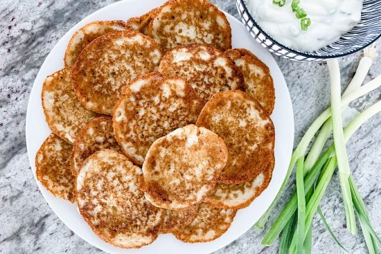 Fried crispy, tender Polish potato pancakes stack on a white plate. The plate is placed on a grey marble countertop. Beside the plate, on the right top side of the plate, is a small white serving bowl with black stripes filled with sour cream and topped with fresh green onions. Next to the bowl and plate is a bunch of fresh green onions.