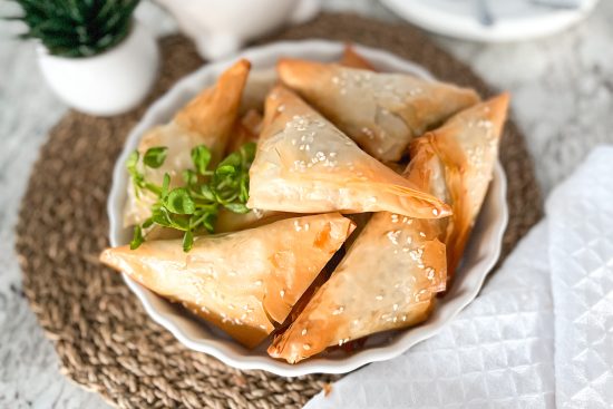 Baked Spinach and goat cheese phyllo triangles and placed on round white porcelain serving plate, with some edible Greens on side. The plate is placed on brown place mat, and beside is placed white kitchen towel. In the background are placed one small white flower pot with green plant and beside white porcelain container.