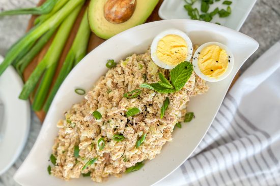 Tuna Egg Salad served in white porcelain oval shaped dish, with one egg cut in half for garnish. The plate is displayed on wood cutting board, white kitchen towel with grey strips, some halves of avocado and green onions beside.