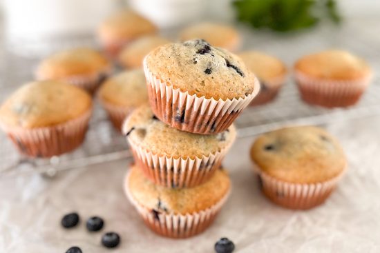 3 Blueberry muffins placed on top of each other. Beside is placed one muffin and some fresh blueberries. In the background some more baked muffins placed on wire cooling rack.