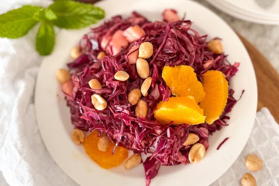 Ready to serve made red cabbage salad on white porcelain serving plate, displayed on grey kitchen counter, beside fresh basil leafy brunch