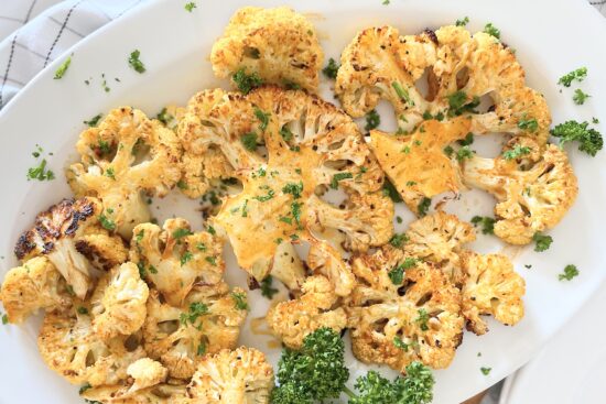 Baked cauliflower, sprinkled with fresh chopped parsley, displayed on white porcelain round serving plate.