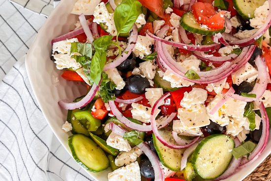Made Greek Salad with Kalamata olives served in white porcelain bowl, beside white kitchen towel with grey strips.