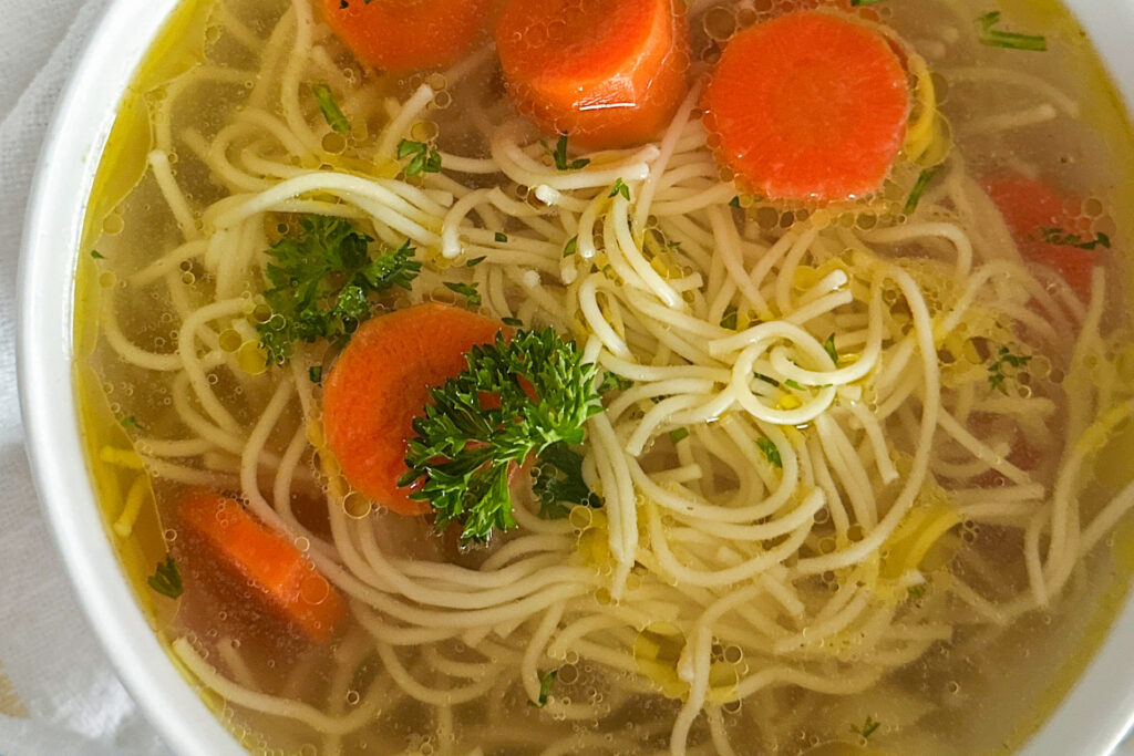 A white serving bowl filled with rosół, a clear Polish chicken soup, featuring tender noodles, sliced carrots, and a sprinkle of fresh parsley as garnish. The bowl is set on a clean white surface.