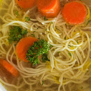 A white serving bowl filled with rosół, a clear Polish chicken soup, featuring tender noodles, sliced carrots, and a sprinkle of fresh parsley as garnish. The bowl is set on a clean white surface.
