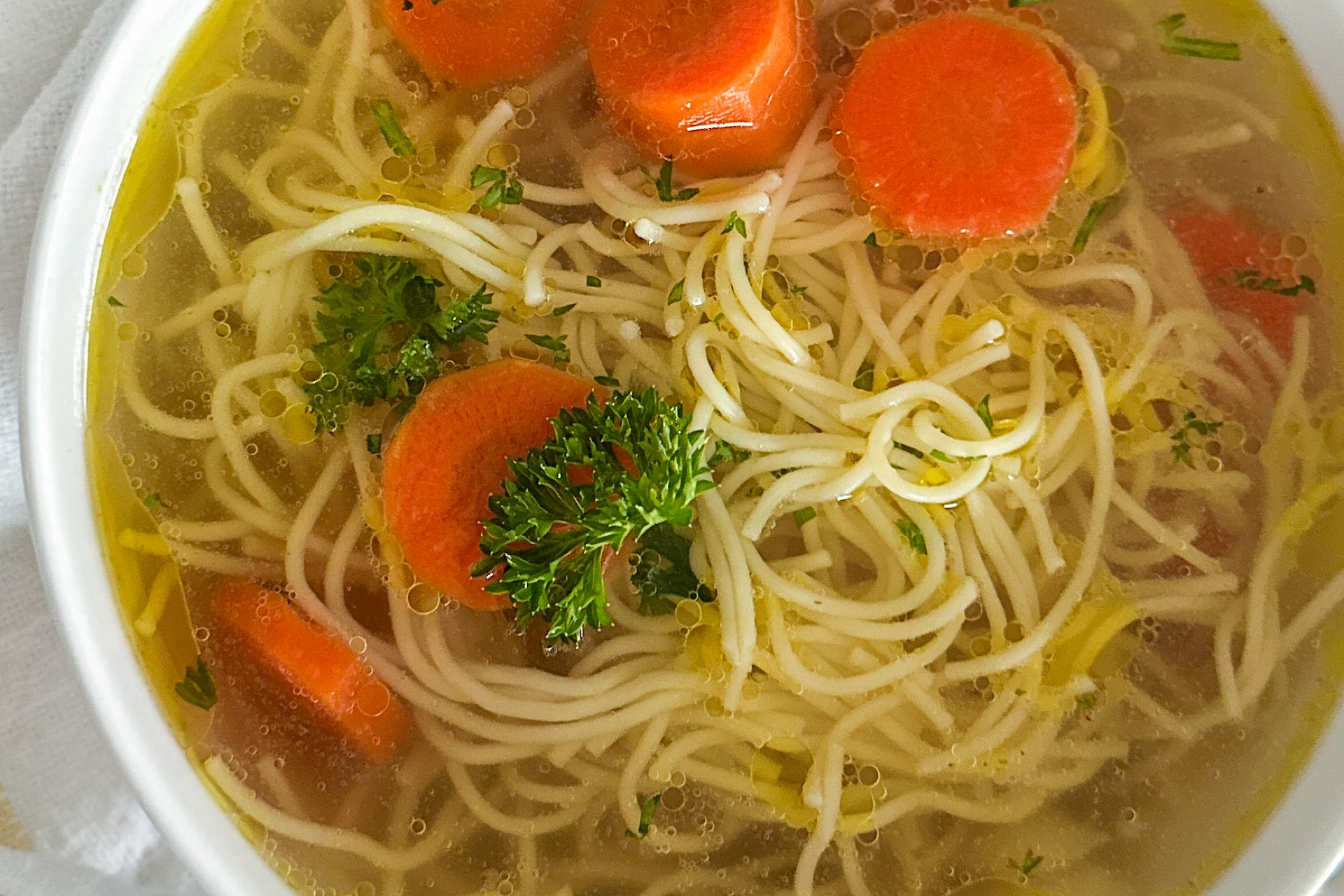 A white serving bowl filled with rosół, a clear Polish chicken soup, featuring tender noodles, sliced carrots, and a sprinkle of fresh parsley as garnish. The bowl is set on a clean white surface.
