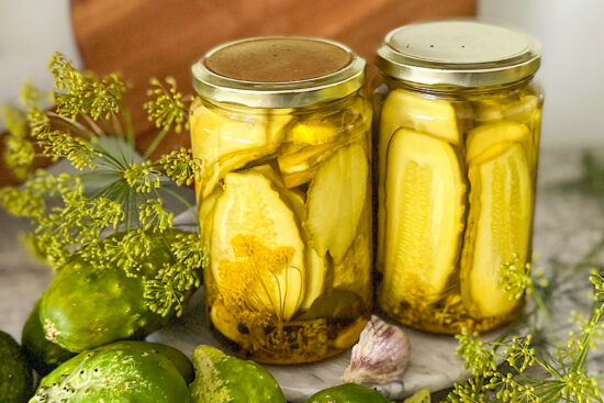 Canning jars filled with curry pickles, beside small pickling cucumbers, fresh dill heads and clove of garlic.