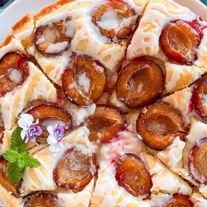 Baked plum cake on white plate, placed on blue kitchen towel, beside few purple plums and fresh mint leaves.