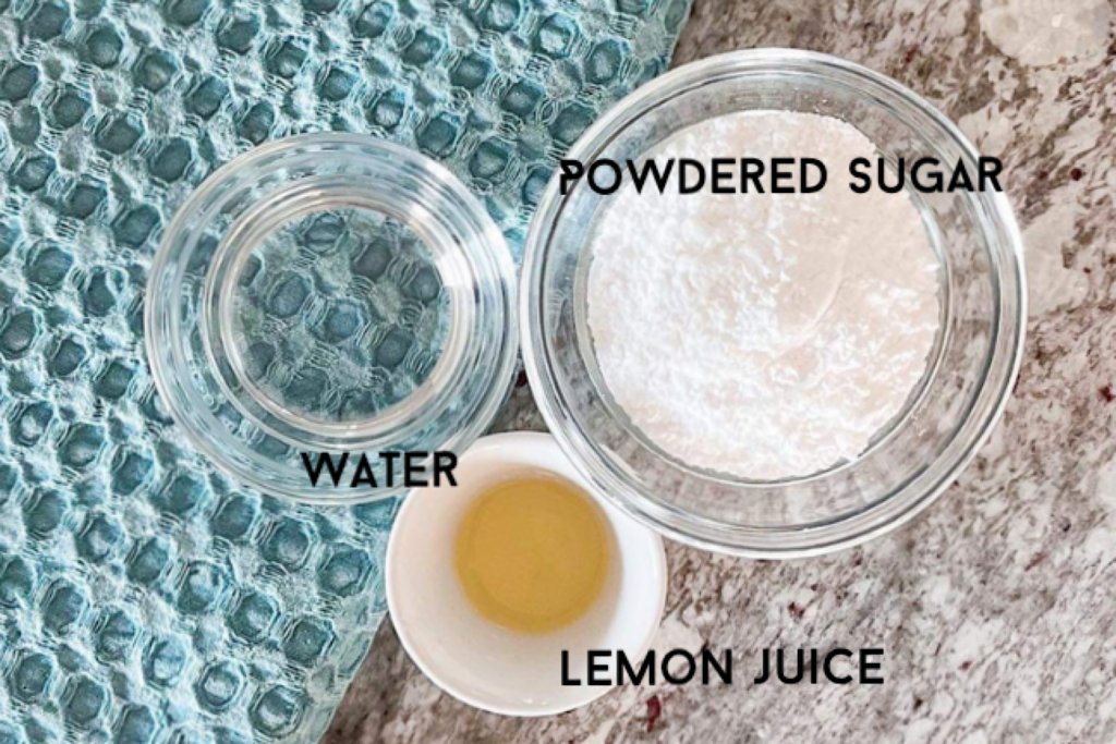 In glass bow: powdered sugar, lemon juice and water, beside blue kitchen towel placed on grey counter top.