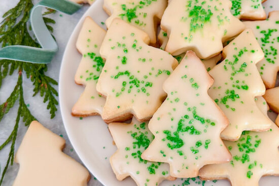 Baked Christmas Tree shaped Orange Sugar cookies topped with orange glaze and sprinkled with green sugar sprinkles, displayed on white ceramic plate. Beside some more sugar cookies and fresh Christmas greenery and green ribbon.