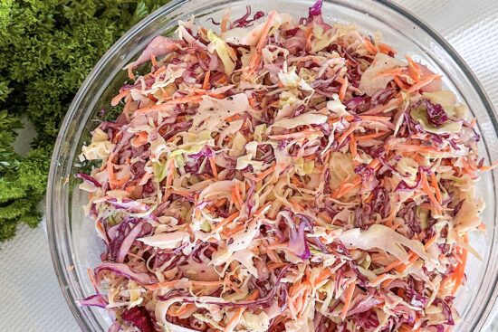 Coleslaw Recipe Salad in glass serving bowl, beside fresh green Parsley bunch, displayed on white kitchen cloth.