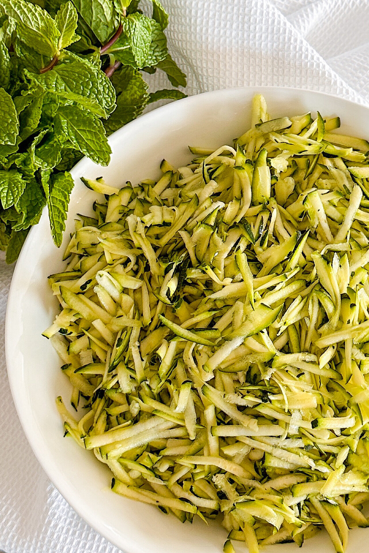 Shredded zucchini in white porcelain bowl beside bunch of fresh green mint.