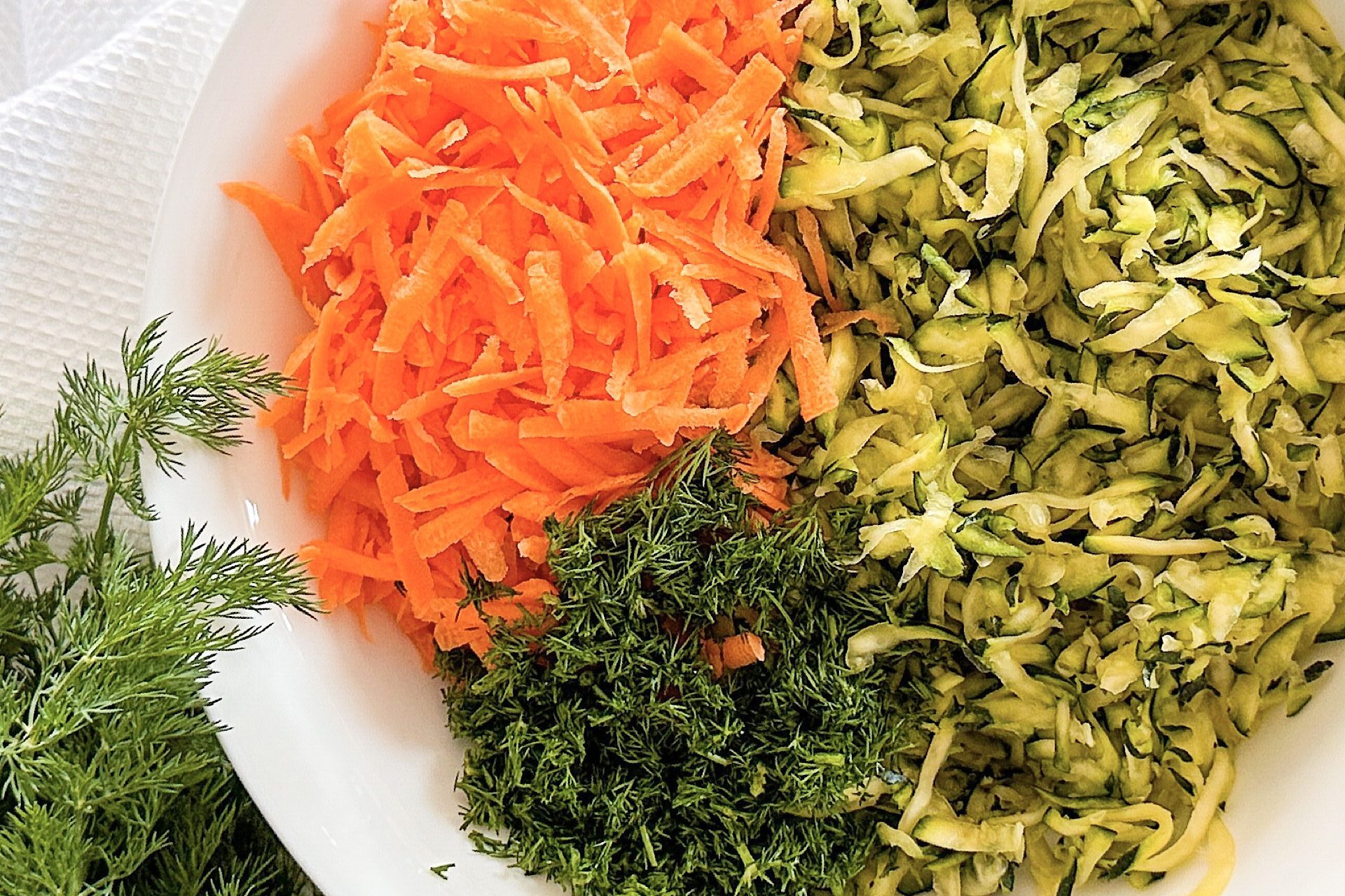 Shredded zucchini, carrot, and chopped dill in white plate, beside the plate bunch of fresh mint.