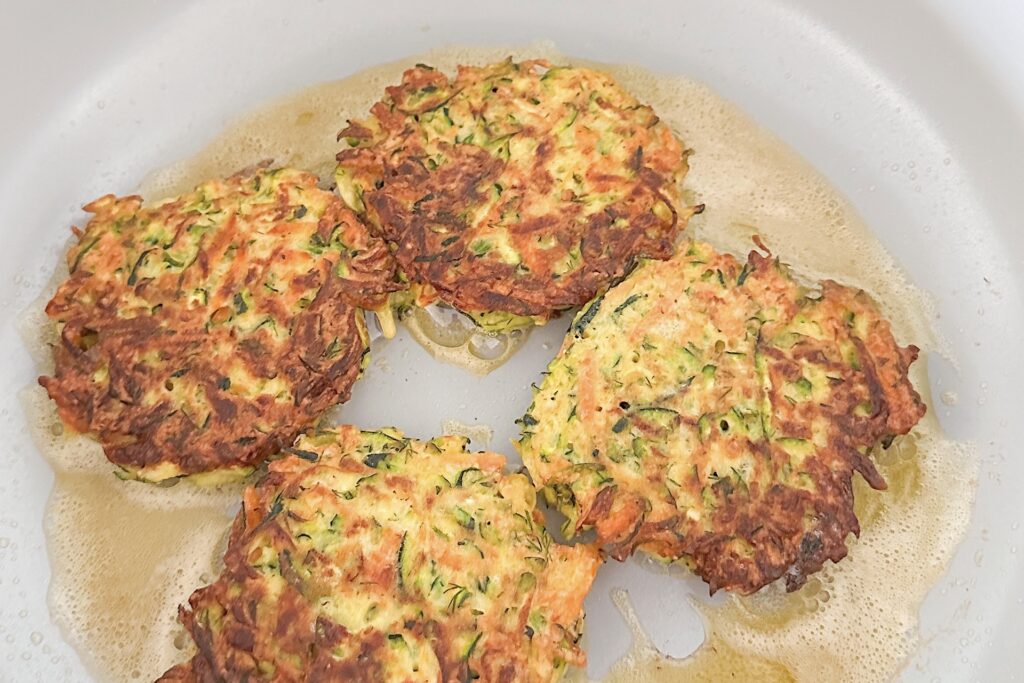 Golden 4 zucchini fritters frying on light grey stove pan with oil.