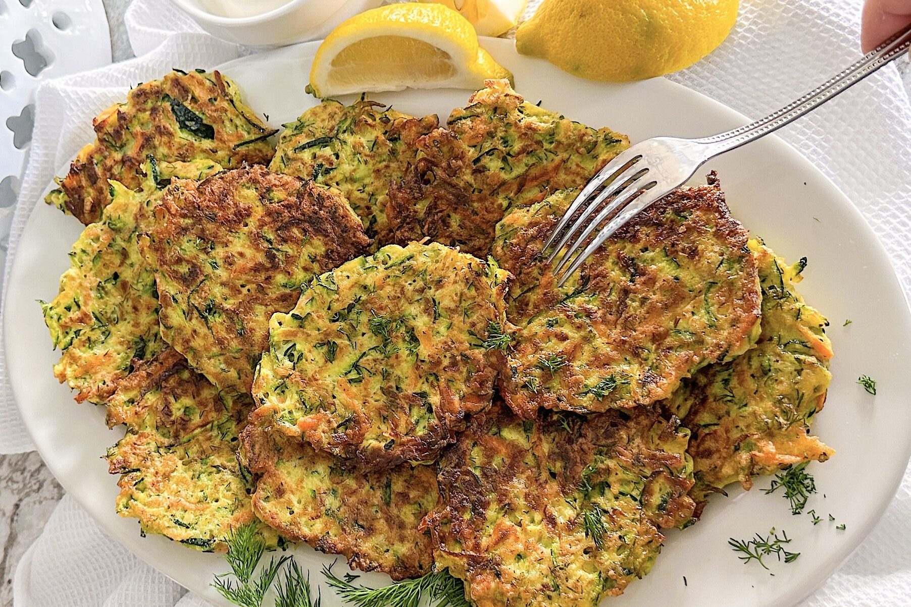 11 fried zucchini fritters displayed on oval white porcelain plate. Beside, 2 pieces of Lemon, hand holding fork that is touching one fritter. Plate is displayed on white kitchen towel over the grey kitchen countertop.