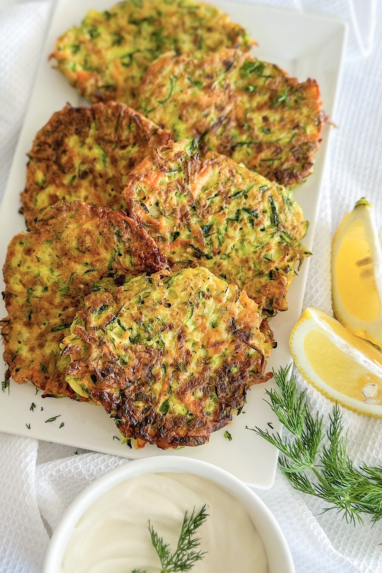 Fried 6 zucchini carrot fritters displayed on white porcelain serving plate, Sprinkle with fresh chopped dill, beside 2 pieces fresh lemon, small bowl filled with yogurt and one small piece of fresh dill. Plate is laying down on white kitchen towel.