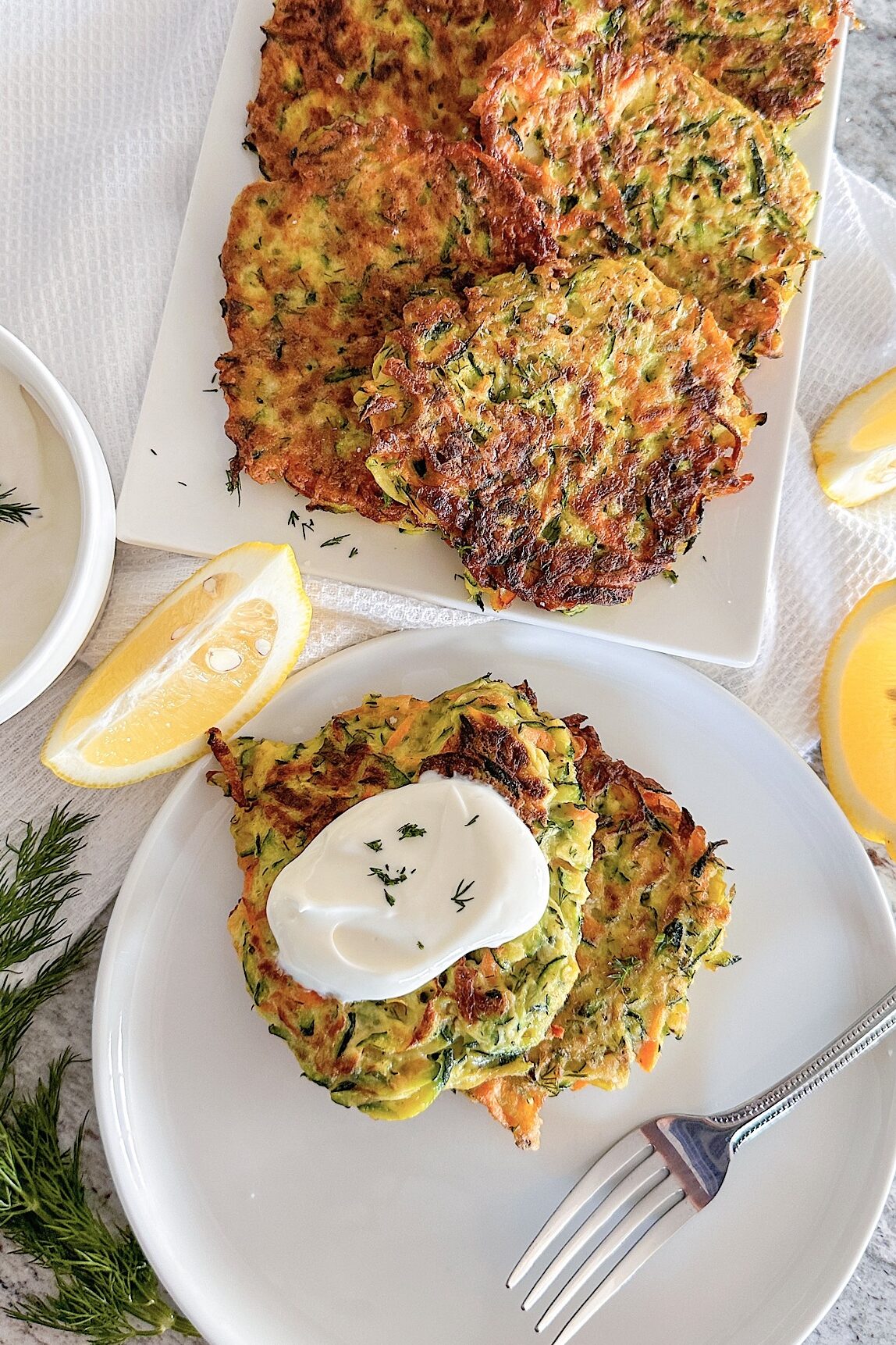 Fried 6 zucchini carrot fritters displayed on white porcelain serving plate, Sprinkle with fresh chopped dill, beside 3 pieces fresh lemon, small bowl filled with yogurt and one small piece of fresh dill, and one round white plate with two fritters topped with spoon of sour cream, on that plate also is placed fork. Plate is laying down on white kitchen towel.