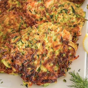 Fried 4 zucchini carrot fritters displayed on white porcelain serving plate, Sprinkle with fresh chopped dill, beside 2 pieces fresh lemon. Plate is laying down on white kitchen towel.
