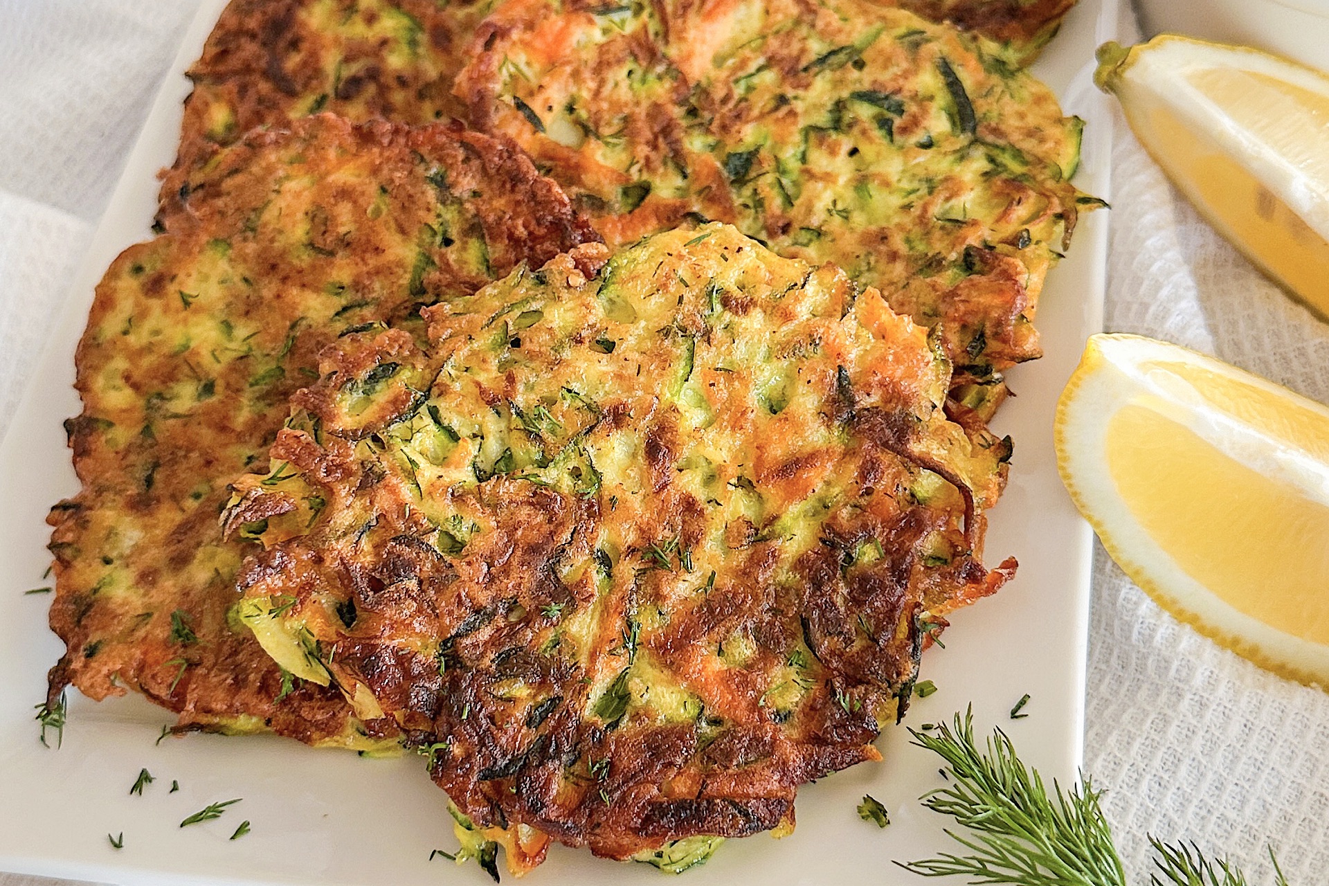 Fried 4 zucchini carrot fritters displayed on white porcelain serving plate, Sprinkle with fresh chopped dill, beside 2 pieces fresh lemon. Plate is laying down on white kitchen towel.