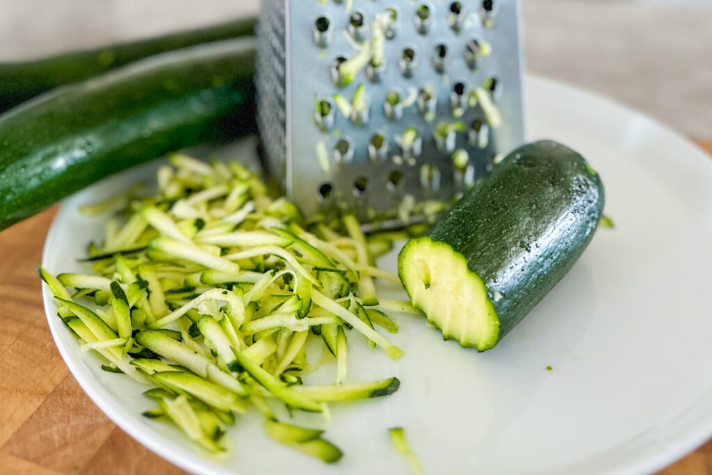 On white plate is placed manual grater, beside some shredded zucchini with half of the zucchini and two zucchini's in the background. The plate is place on brown wood cutting board. 