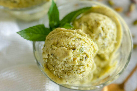 2 scoops of No churn Pistachio Ice Cream in glass serving bowl with some fresh mint on as a garnish. Glass bowl is standing on white kitchen towel.