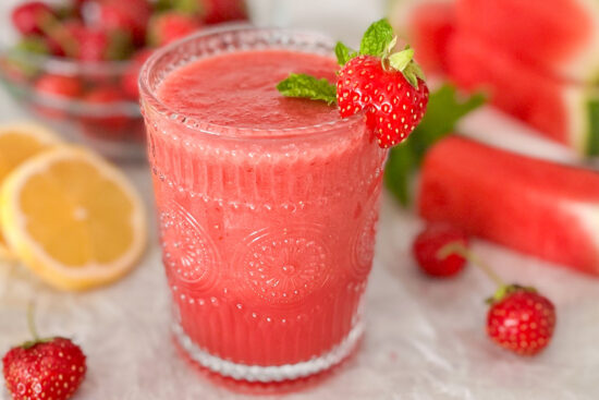 Drinking glass filled with strawberry watermelon smoothie. On the edge of the glass is placed one small strawberry. The glass is standing on white surface, beside the glass are some pieces of cut watermelon, fresh green mint and two lemon slices. In the background are some strawberries in glass bowl.