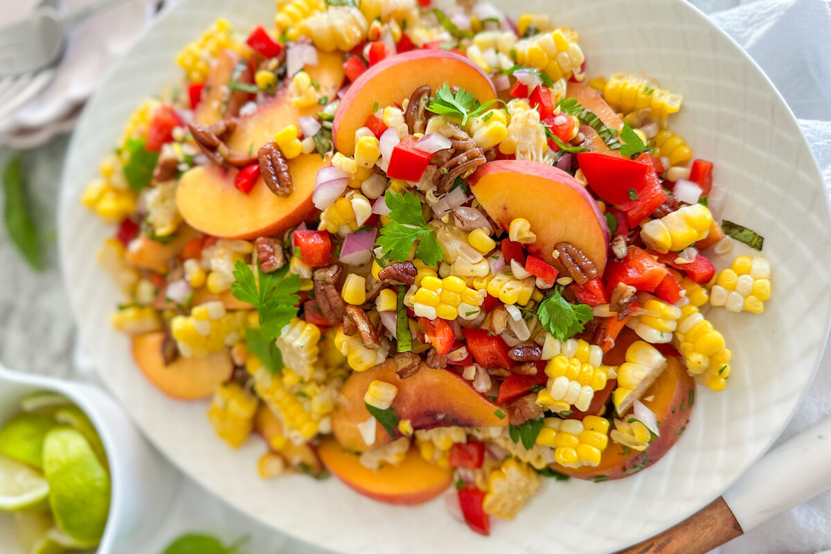 Sweet Corn Peach salad displayed on oval white serving plate. In left down corner is small bowl filled with sliced limes.