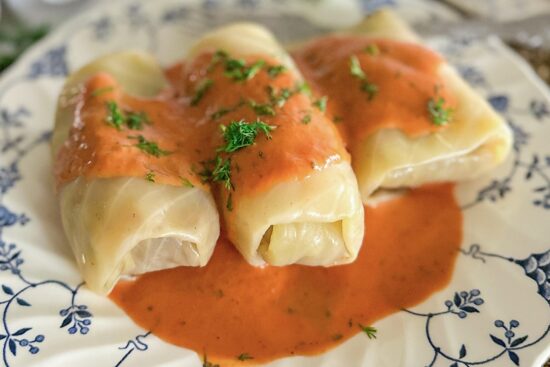 Three cooked, stuffed cabbage rolls are placed on a white plate with dark blue floral designs. The rolls are topped with tomato sauce and garnished with freshly chopped dill.