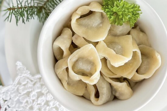 A white porcelain serving dish filled with cooked mushroom dumplings is placed on a large white porcelain plate. A small piece of fresh parsley garnishes the dish. In the top right corner, a small part of a fresh Christmas tree branch is visible, while in the bottom left corner, small star-shaped crochet doilie is placed.