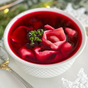 A serving bowl filled with cooked mushroom dumplings and beetroot broth ('borsch') is placed on a large white porcelain serving plate. A large silver spoon rests on the left side, while a fresh Christmas tree branch with some light twinkle lights adds decoration on top. Small star-shaped doilies are placed on the top right and bottom right corners.