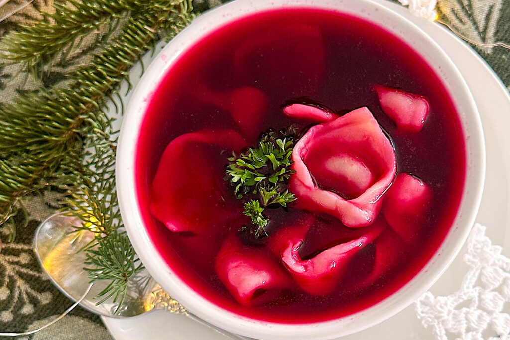 A serving bowl filled with cooked mushroom dumplings and beetroot broth ('borsch') is placed on a large white porcelain serving plate. A large silver spoon rests on the left side, while a fresh Christmas tree branch with some light twinkle lights adds decoration on top. Small star-shaped doilies are placed on the top right and bottom right corners.