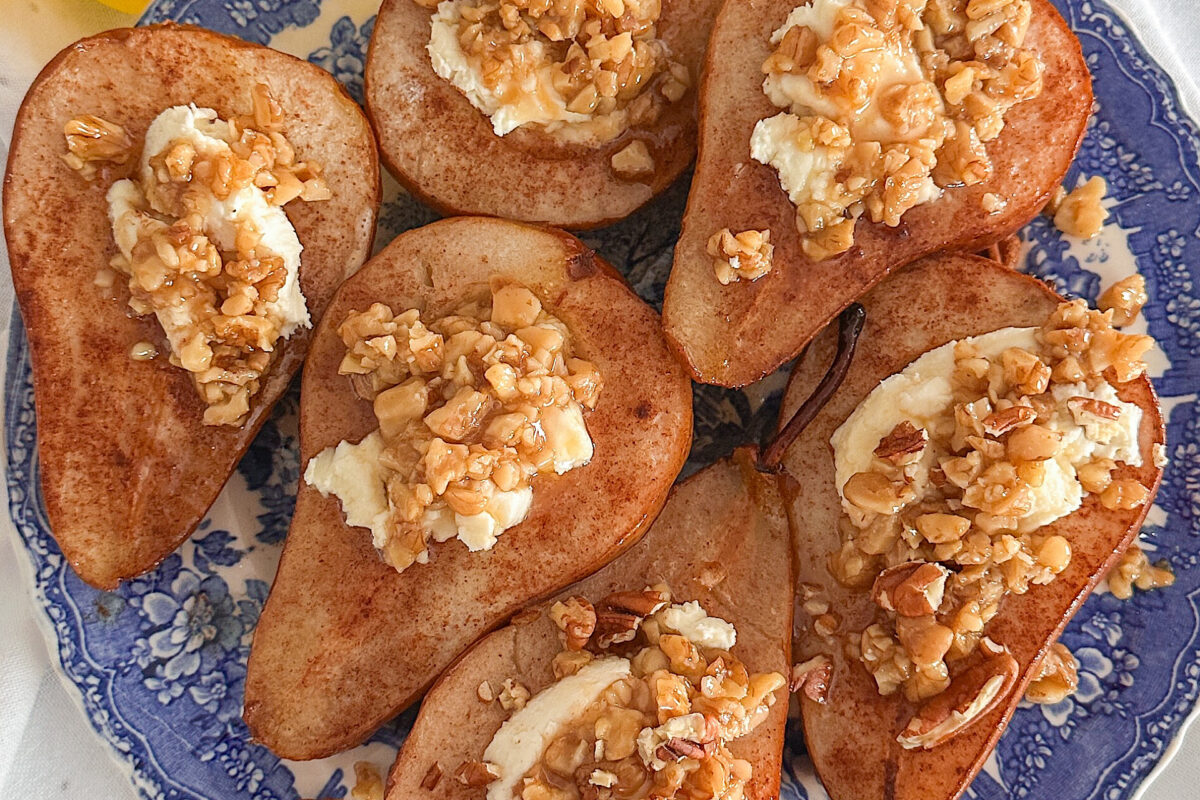 Baked pears, sprinkle with cinnamon and stuffed with goat cheese and topped with honey-butter-roasted pecans are placed on a porcelain plate with a royal blue design. The plate rests on a white kitchen towel.
