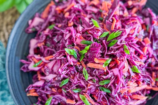 The already prepared and mixed red cabbage coleslaw is placed in a black serving bowl and placed on a brown wooden serving mat. In the bottom left corner, a light blue kitchen towel is visible, and in the top left corner, a few fresh mint leaves are scattered.