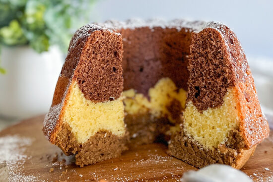 The Tiramisu Bundt Cake is sliced to reveal its three distinct layers and placed on a round brown cutting board. In front of the board, a few slices of the cake are arranged alongside a powdered sugar shaker. In the background, on the right side, there's a white flower pot containing green leaves.