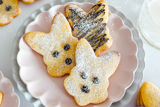 Three baked Easter Bunny Cookies are displayed on a pink porcelain serving plate, which is placed on top of a light grey serving plate. The plates sit on a white surface. In the top right corner, there is a glass filled with milk. In the bottom right corner, a small part of a stag cookie with a purple ribbon is visible. In the top left corner, a few more baked cookies can be seen, and in the bottom left corner, and small part of a pink plate with cookie is visible, and powdered sugar shaker.