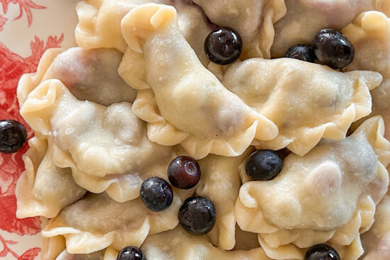 A bunch of cooked soft, Blueberry pierogi are placed on a white plate with a red flower design. Few fresh blueberries are placed on the top of pierogi.
